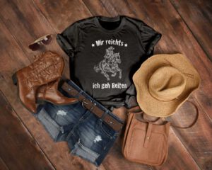 Ein graues T-Shirt mit dem Aufdruck „Mir reicht’s, ich geh reiten!“ liegt auf einem Holzhintergrund, daneben ein Cowboyhut, Westernstiefel und ein Gürtel – perfekt für Fans des Westernreitens mit Sinn für Humor.